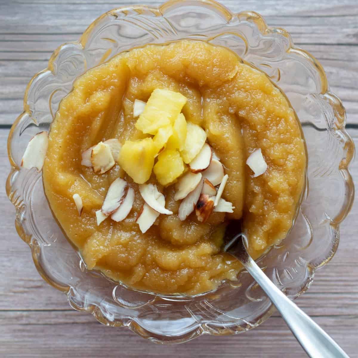 Almond pineapple halwa served in a glass bowl with a dessert spoon and pineapple pieces and almond pieces garnished on top