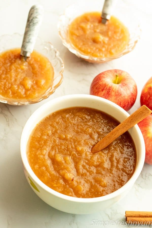 Instant pot apple sauce ready to serve in a white bowl with cinnamon stick dipped in it and also served in two glass bowls beside it with apples on the side.
