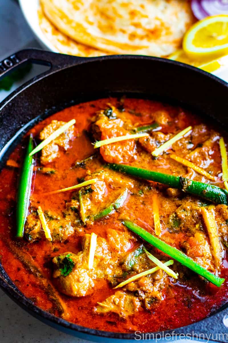 Close up image of Chicken Karahi in a black cast iron wok garnished with thin long slices of jalapenos and ginger juliennes with a plate of paratha and lemon slices on the side.