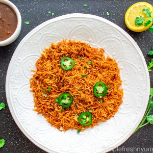 Arroz Rojo/Spanish rice served in a round white plate with jalapeno slices and cilantro garnished on top. There is a lemon on one side and a bowl of black bean soup on another side.