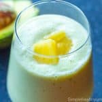 Close up image of Avocado pineapple smoothie in a glass with pineapple pieces garnished on top and a half avocado on the side.