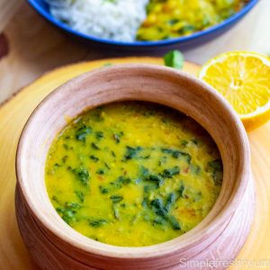 Spinach Dal in a pottery serving dish with a half lemon on the side and a serving plate with dal and white rice in the background.