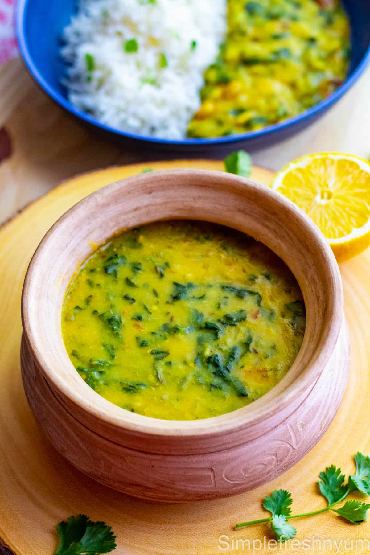 Spinach dal served in a light brown color pottery dish with a slice of lemon on the side and a blue plate of spinach dal served with white rice in the back ground.