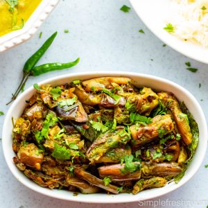 Brinjal fry in a white oval serving dish with two thai green chillies on the side and cilantro garnished on top
