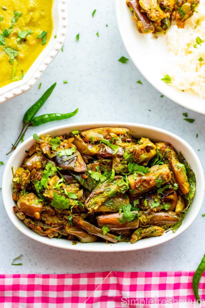 Brinjal fry served in a white oval serving dish with a dish of dal on the side and a plate served with rice, brinjal fry and dal. Cilantro is garnished on top.
