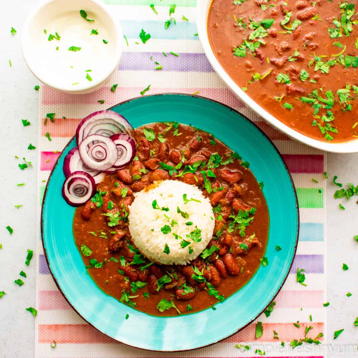 Rajma served in a blue plate with white rice and raw onion rings. There is a bowl of yogurt on the side and a white serving bowl full of Rajma. Cilantro is garnished on top