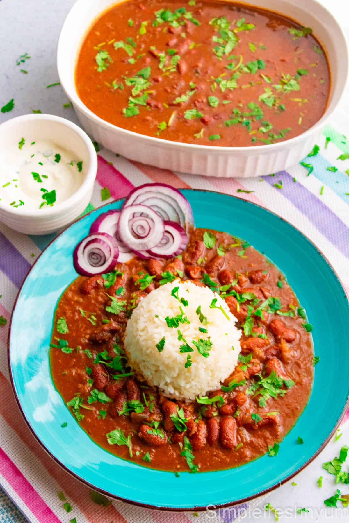 Indian style kidney bean curry Rajma served in a blue plate with white rice and yogurt on the side with cilantro on top. A serving bowl full of Rajma is also there on the side.