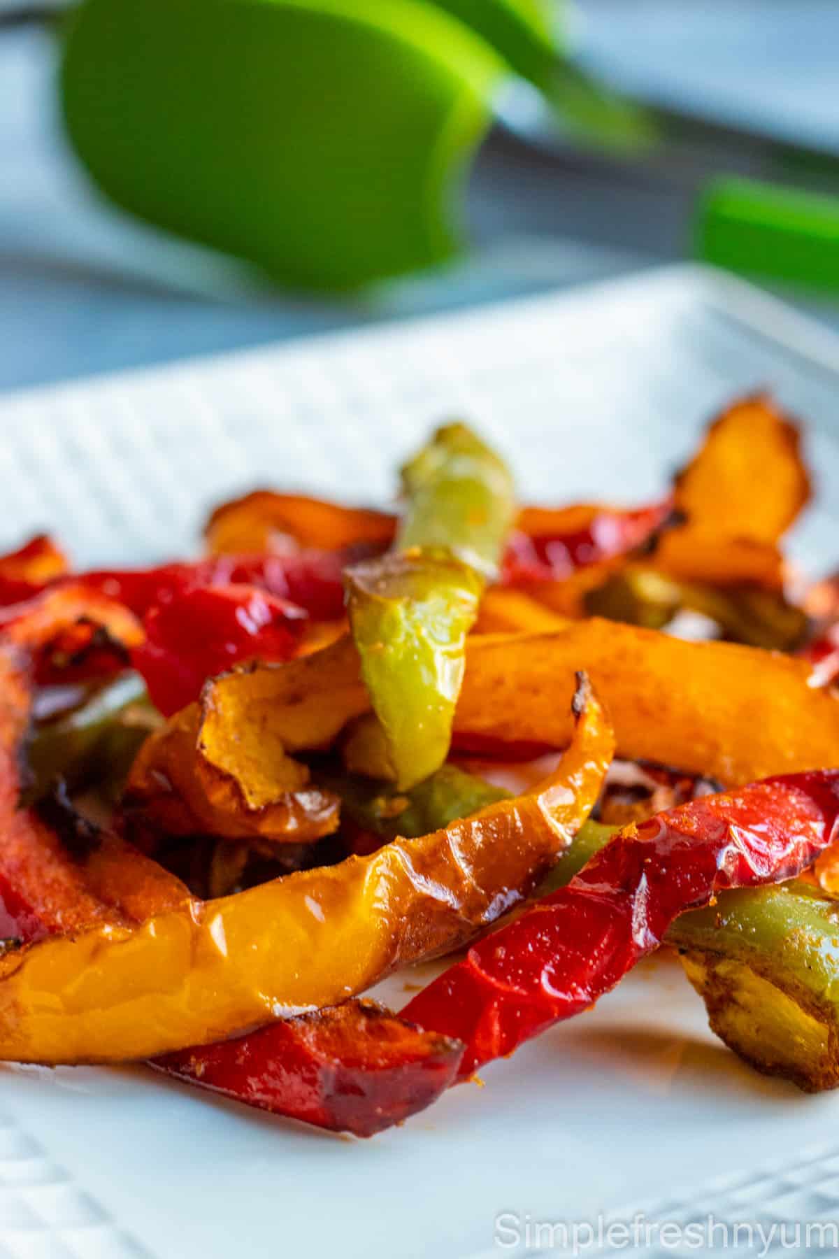Close up image of bell pepper strips roasted in an air fryer served on a white plate with a green tongs in the back ground.