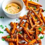 Carrot fries served in a white oval plate with parmesan cheese and cilantro sprinkled on top. Sriracha greek yogurt dip is served in a white round bowl on the side