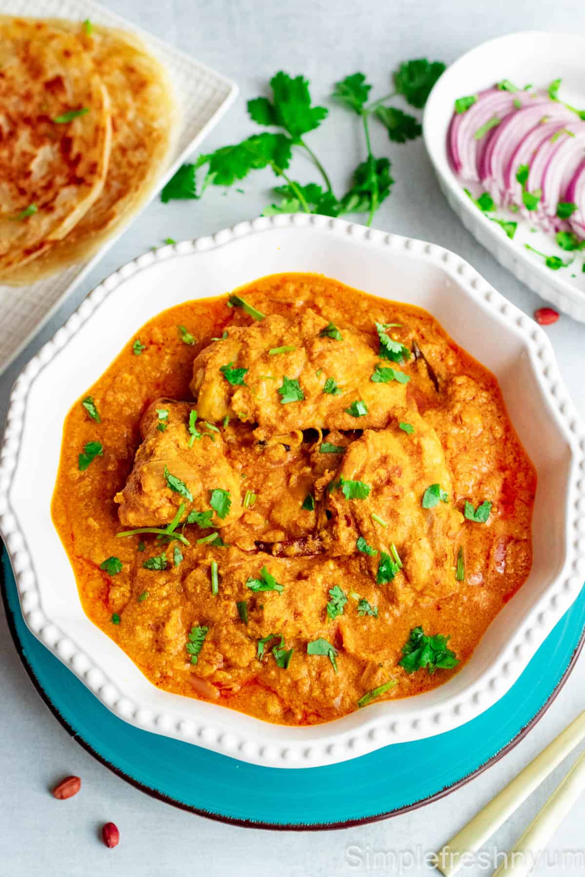Chicken salan curry served in a white round bowl placed on a bluce plate with cilantro garnished on top. A plate of parotta and another serving dish with sliced onions and lemons are placed on the side.