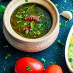 Tomato Rasam served in a clay pot on a black surface with cilantro and red chili garnished on top. There are two roma tomatoes placed on the side of the pot and a serving dish with pongal. Cilantro, garlic cloves and Curry leaves are strewn around the pot for decoration.
