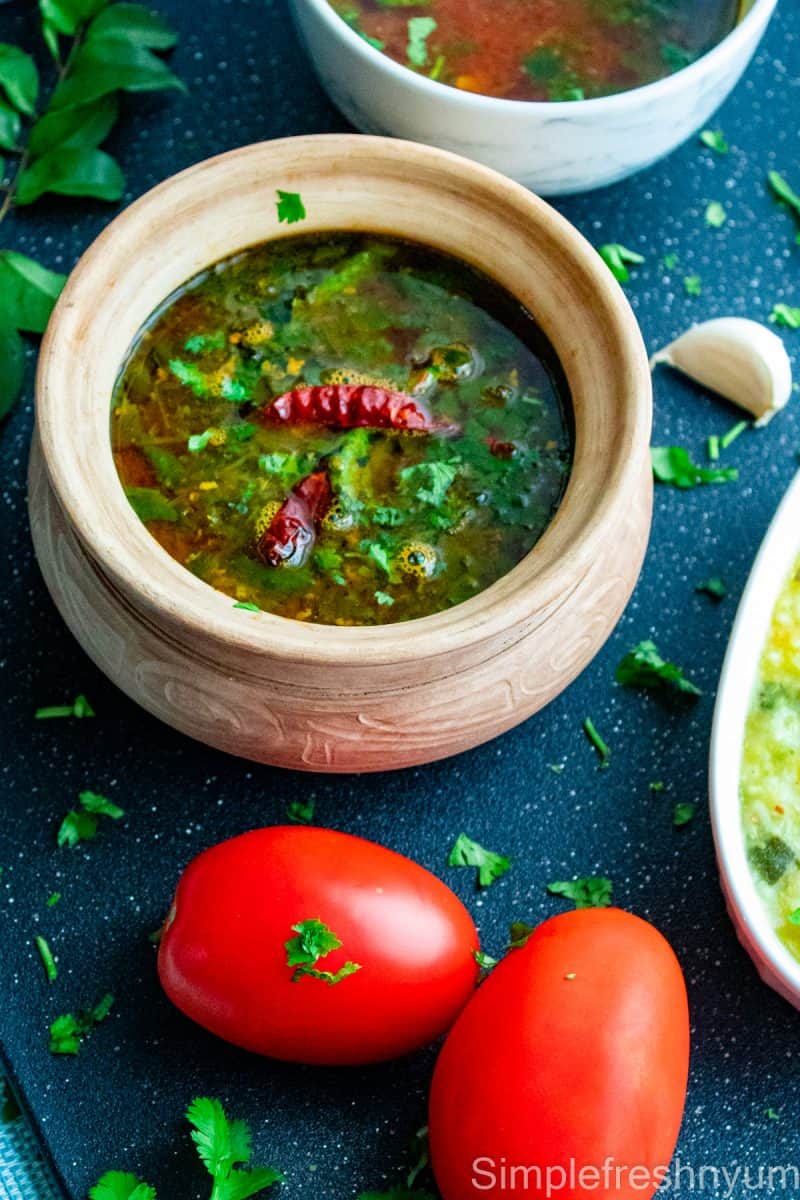 Tomato Rasam served in a clay pot on a black surface with cilantro and red chili garnished on top. There are two roma tomatoes placed on the side of the pot and a serving dish with pongal. Cilantro, garlic cloves and Curry leaves are strewn around the pot for decoration.