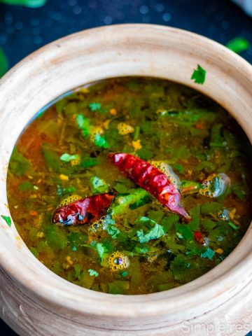 Tomato Rasam served in a light wooden colored clay serving pot with cilantro and red chilies garnished on top.